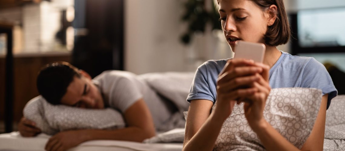 Young woman is secretly sending text messages over smart phone and cheating on her boyfriend who is sleeping on the bed.